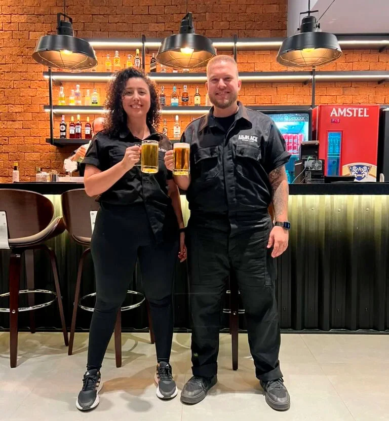 Brenno e Lorena, donos da All Black, de uniforme preto e com dois chopes na bancada do bar